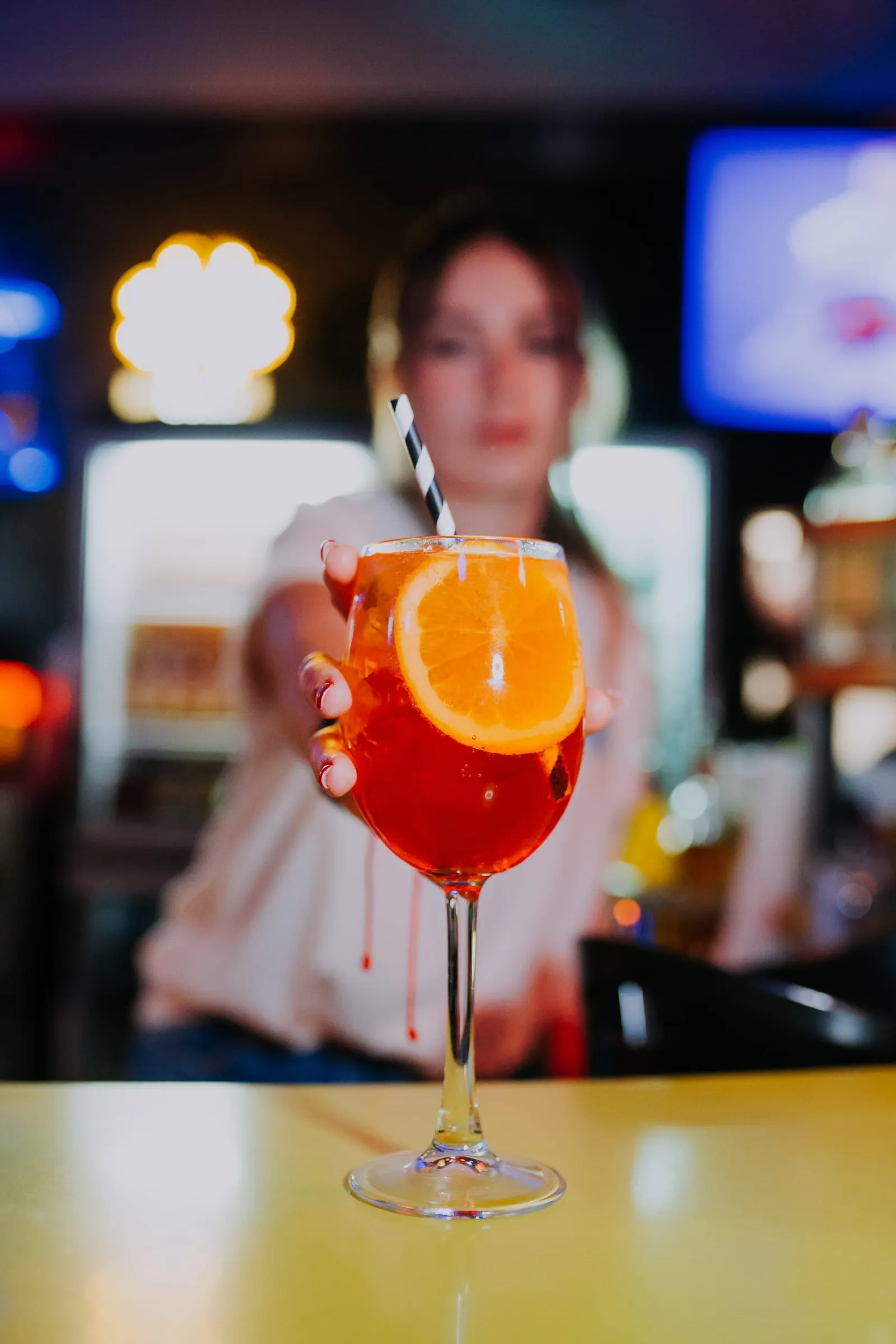 Una camarera de Bulebu acerca a la cámara un cóctel con una rodaja de naranja; detrás, el bar encendido y desenfocado.