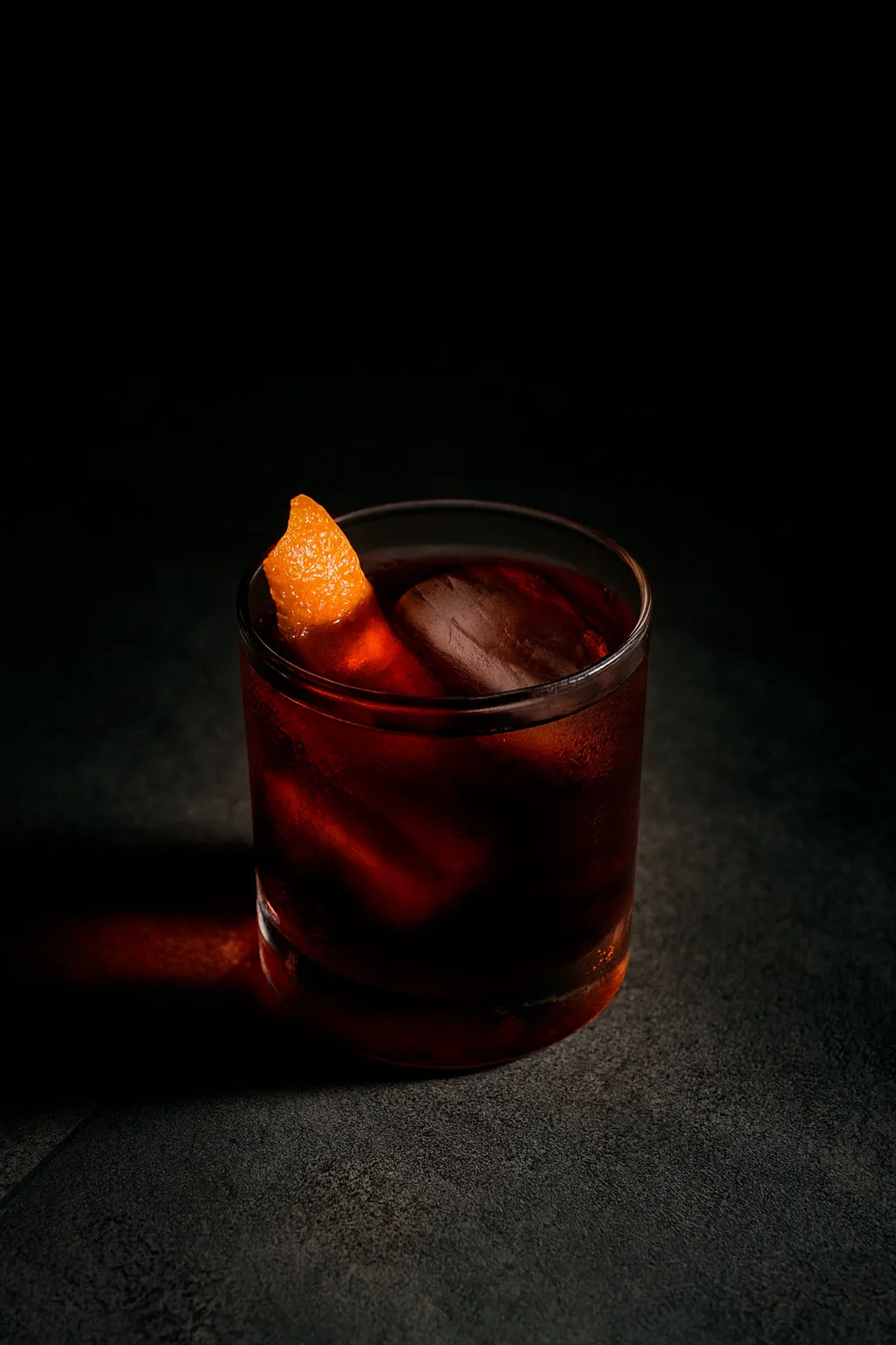Cóctel rojizo con hielo y piel de naranja, iluminado sobre fondo oscuro.